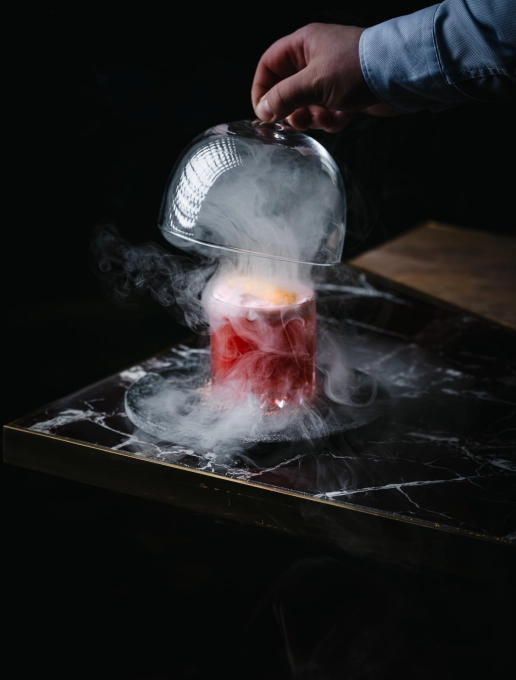 Cocktail rouge où une main retire une cloche en verre pour laisser s’échapper la fumée de la boisson