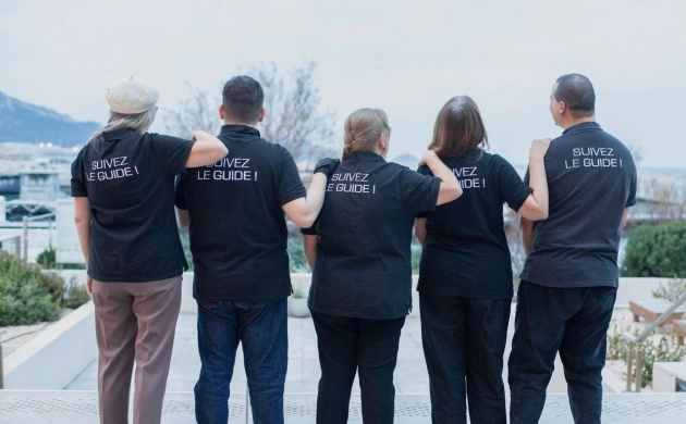 Cinq personnes sont de dos et portent le même tee-shirt noir où il est inscrit « Suivez le guide ! »