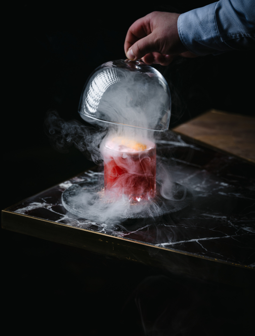 Cocktail rouge où une main retire une cloche en verre pour laisser s’échapper la fumée de la boisson