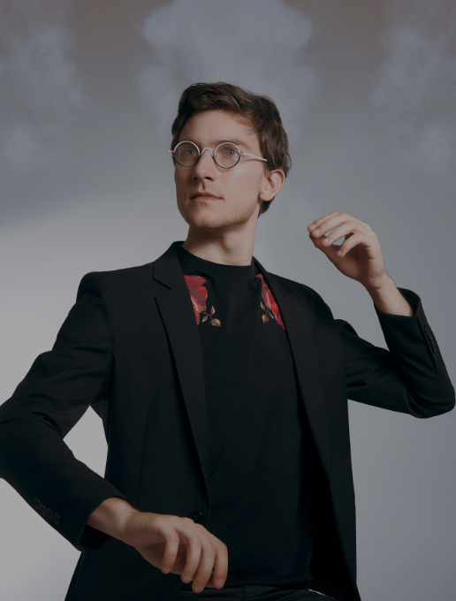 Portrait de Pierre Fontenelle, très élégant en costume noir, portant des lunettes rondes, sur un fond gris avec un effet de fumée en arrière-plan.