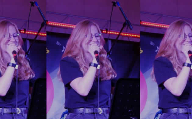 Nina, jeune musicienne, chante sur scène, un micro à la main, sur un fond tamisé éclairée par des néons violet et rouge